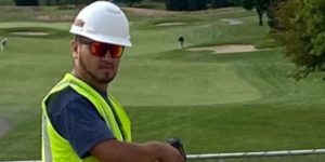 tradesman with the drill motor, hammer, hard hat, and golf course in the background